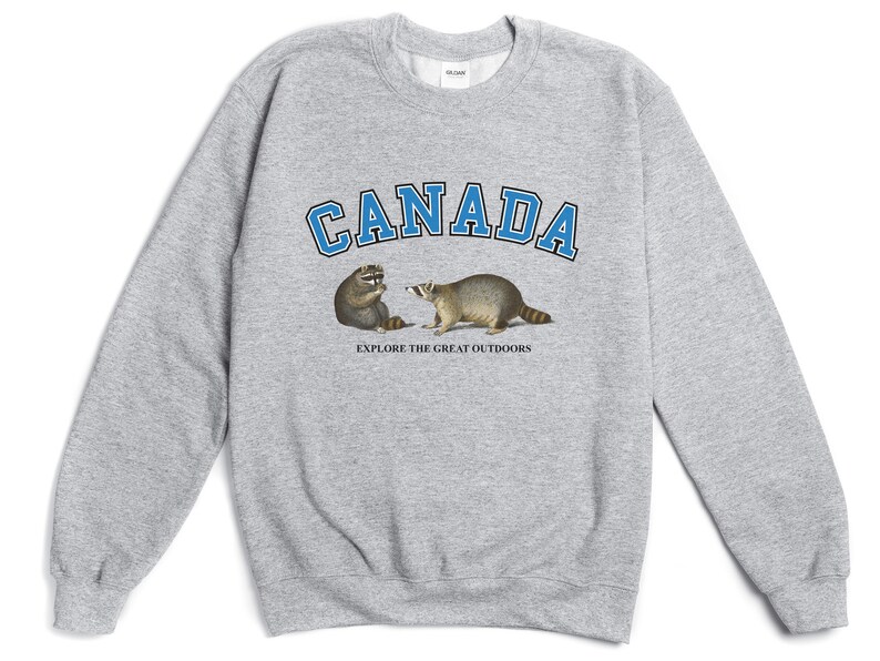 May include: A heather gray crewneck sweatshirt featuring the word CANADA in blue block letters with black outlines. Below the text are two raccoons and the phrase EXPLORE THE GREAT OUTDOORS printed in small black capital letters on the chest.
