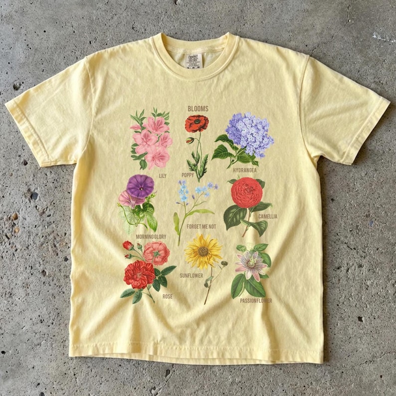 May include: A light yellow t-shirt featuring a floral design with the word "Blooms" at the top. The design includes illustrations of various flowers such as lilies, poppies, hydrangeas, and roses. The shirt is made of a soft, comfortable material.