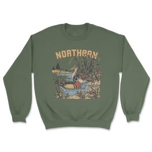 May include: An olive green crewneck sweatshirt featuring a graphic illustration of two ducks swimming in a pond surrounded by reeds and cattails. The text NORTHERN is printed in a tan, arched font above the detailed nature scene.