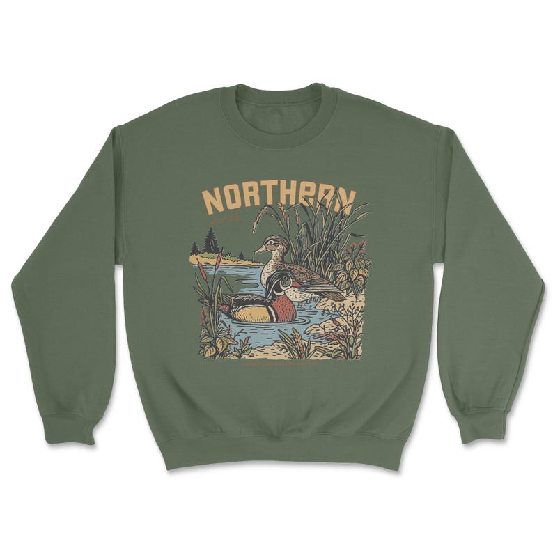 May include: An olive green crewneck sweatshirt featuring a graphic illustration of two ducks swimming in a pond surrounded by reeds and cattails. The text NORTHERN is printed in a tan, arched font above the detailed nature scene.
