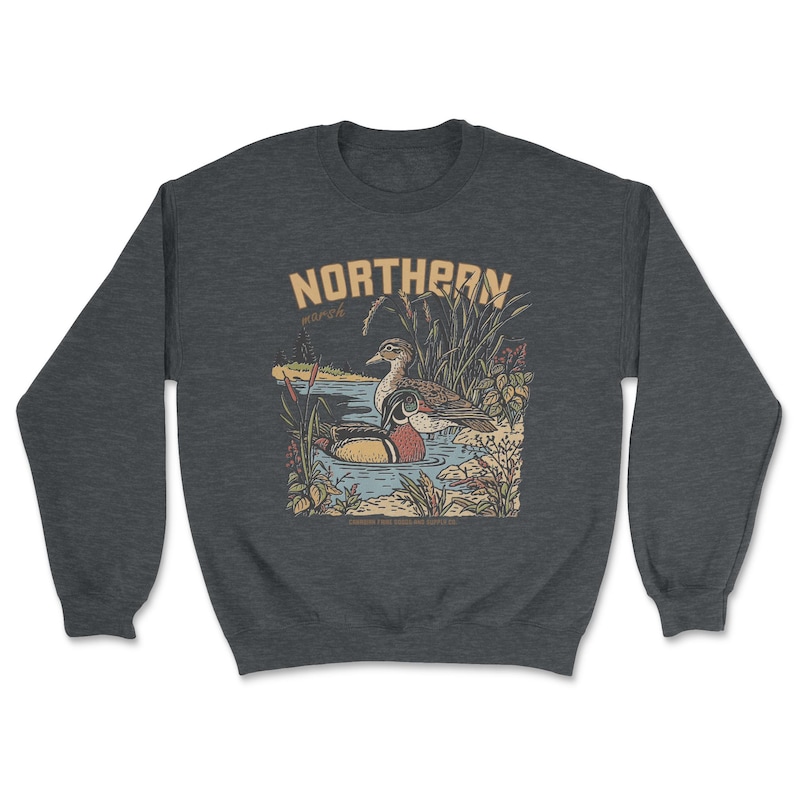 May include: A dark heather gray crewneck sweatshirt featuring a graphic illustration of two ducks swimming in a pond surrounded by reeds and plants. The text NORTHERN is printed in large tan letters above the ducks, with the word march below it.