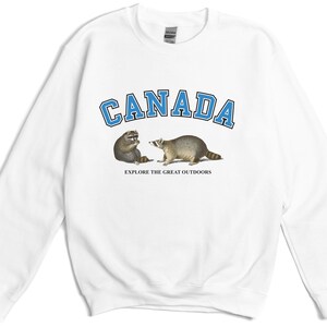 May include: A white long-sleeve sweatshirt featuring the word CANADA in blue block letters with a black outline. Below the text are two raccoons facing each other, with the phrase EXPLORE THE GREAT OUTDOORS printed in small black capital letters.
