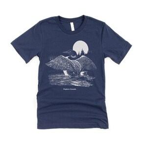 May include: A navy blue t-shirt featuring a white graphic of a loon with its wings spread, standing in a lake under a full moon. The text "Explore Canada" is printed below the loon.