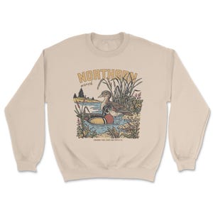 May include: A beige crewneck sweatshirt featuring a graphic illustration of two ducks in a marsh. The text NORTHERN is printed in large letters above the ducks, with marsh below it. Small text at the bottom reads CANADIAN FIBRE GOODS AND SUPPLY CO.