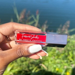 Könnte beinhalten: Eine durchsichtige Glasflasche mit einer roten Flüssigkeit. Die Flasche hat einen silbernen Verschluss und ein weißes Etikett mit dem Text "Forever Shades By Jamaria Chenelle".