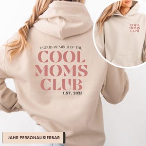 Könnte beinhalten: Beige Kapuzenpullover mit dem Text "COOL MOMS CLUB" in einem rosaroten Farbton auf der Rückseite und der Vorderseite. Der Hoodie hat auch den Text "EST. 2025". Der Hoodie hat eine Kapuze und eine Vordertasche.