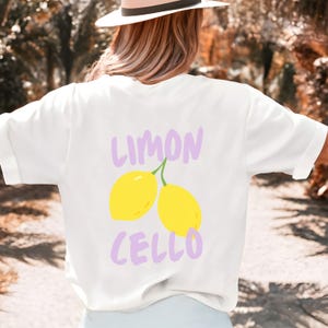 K&ouml;nnte beinhalten: Wei&szlig;es T-Shirt mit einem "LIMON CELLO" Design in Lavendel und Gelb. Das Design zeigt zwei gelbe Zitronen mit gr&uuml;nen Stielen. Das Shirt ist ein l&auml;ssiger Stil mit kurzen &Auml;rmeln.
