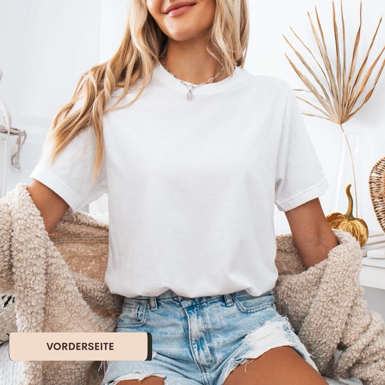 K&ouml;nnte beinhalten: Ein wei&szlig;es T-Shirt mit Rundhalsausschnitt und kurzen &Auml;rmeln. Dazu werden Jeansshorts und eine beige Strickjacke getragen. Der Text "VORDERSEITE" ist auf einem rechteckigen Etikett zu sehen. Die Person tr&auml;gt eine silberne Halskette.