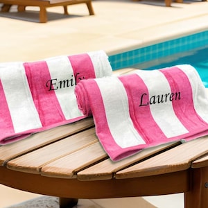 May include: Two rolled beach towels with pink and white stripes rest on a wooden table. The towels are personalized with the names "Emily" and "Lauren" in black script. The scene is set near a pool.