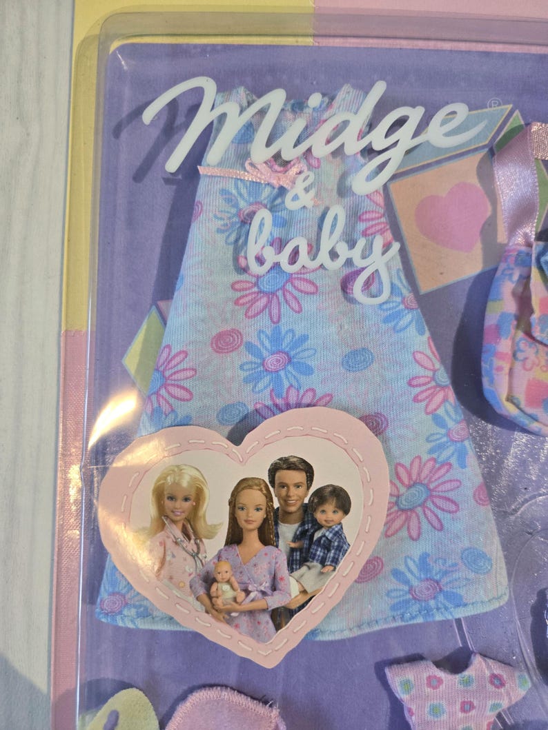 May include: A Midge & baby doll set in its original packaging. The packaging features a light blue dress with pink and blue floral patterns. A heart-shaped image shows a family of dolls. The text "Midge & baby" is visible.