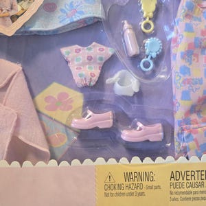 May include: A baby doll clothing set in a clear plastic package. The set includes a pink blanket, a patterned shirt and pants, a pair of pink shoes, a bottle, and a rattle. The warning label states "Choking Hazard - Small parts."