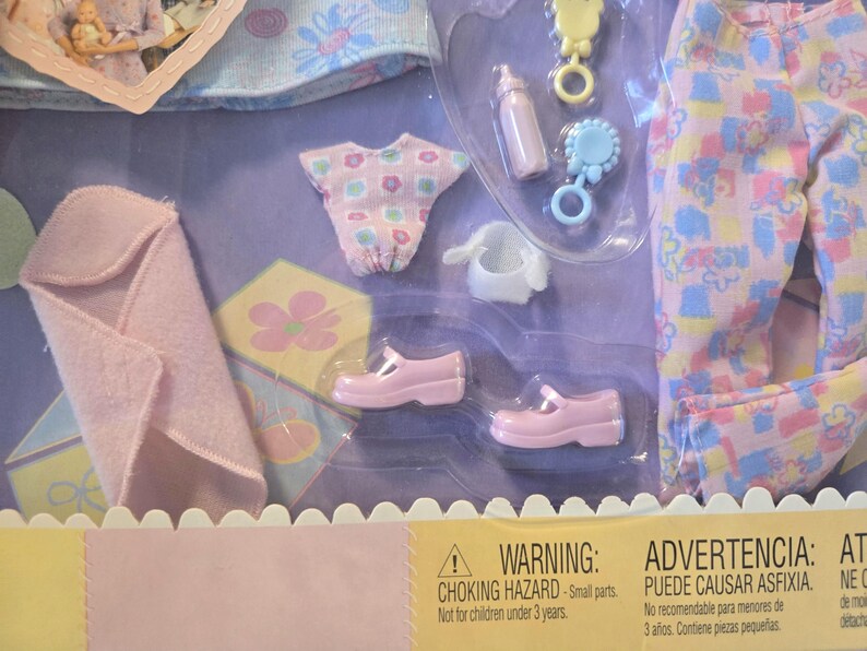 May include: A baby doll clothing set in a clear plastic package. The set includes a pink blanket, a patterned shirt and pants, a pair of pink shoes, a bottle, and a rattle. The warning label states "Choking Hazard - Small parts."