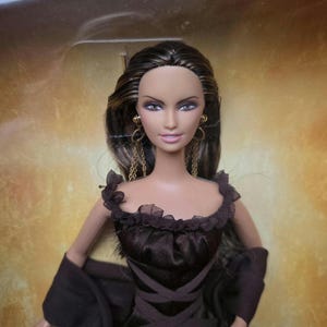 May include: A fashion doll with long, dark hair and blue eyes, wearing a dark brown dress with a ruffled neckline and a crisscross bodice. The doll has gold-colored earrings and a matching shawl.