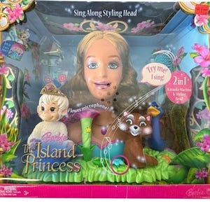 Puede incluir: Una cabeza de peinado Barbie "The Island Princess" en una caja. La caja presenta una cabeza de muñeca, una muñeca pequeña y otras figuras de personajes. La caja tiene el texto "Try me! I sing!" y "2 en 1 Karaoke Machine & Styling Head".