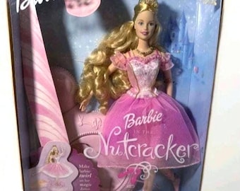 Op de afbeelding: Een Barbie-pop in een doos, getiteld "Barbie in De Notenkraker". De pop draagt een roze tutu en heeft blond haar. De doos bevat de tekst "The Sugarplum Princess" en snoep-thema graphics.