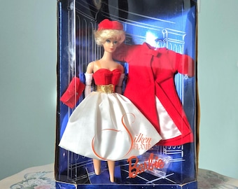 May include: A vintage Barbie doll in a clear plastic box. The doll wears a red and white dress, a red hat, and a red coat. The box reads "1962 Fashion and Doll Reproductions" and "Collector Edition".