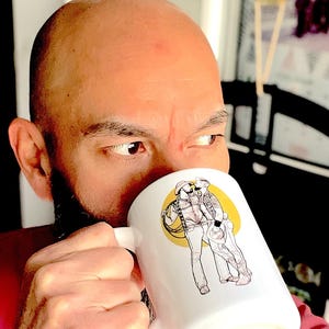 May include: A white ceramic mug with a black and white illustration of two people standing together. The mug has a black handle and is filled with a brown liquid.