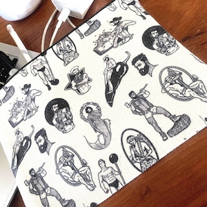 May include: A rectangular, off-white canvas pouch featuring a repeating black and white illustration of various figures. The figures include men in athletic poses, a mermaid, and other stylized characters. The pouch is on a wooden surface.