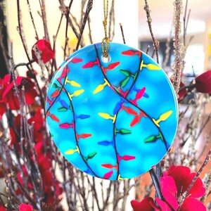 May include: A round, blue Christmas ornament with a colorful string of lights design. The lights are red, yellow, green, and blue. The ornament hangs from a gold string and is surrounded by red flowers and brown branches.