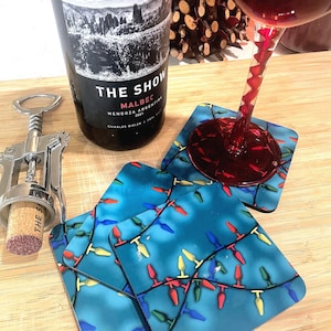 May include: A bottle of Malbec wine, a corkscrew, and four square coasters with a blue background and a pattern of colorful Christmas lights. A glass of red wine is also present.