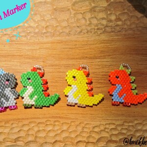 Puede incluir: Cuatro marcadores de punto de dinosaurio de cuentas en diferentes colores: gris, verde, amarillo y naranja. Los marcadores de punto están sobre una superficie de madera. El texto "Stitch Marker" está en la esquina superior izquierda de la imagen.