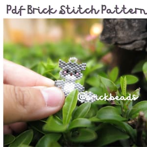 Pode incluir: Um pequeno amuleto de gato de contas cinzas e brancas com olhos cor-de-rosa, segurado na mão contra um fundo de folhas verdes.