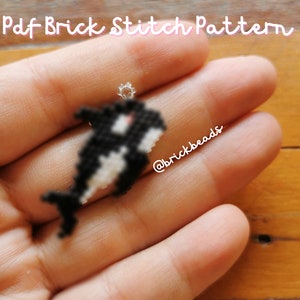 May include: A small, handmade orca charm in a brick stitch pattern. The orca is black and white with a small silver loop at the top. The text "Pdf Brick Stitch Pattern" is above the charm. The charm is held in a hand.