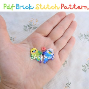 Könnte beinhalten: Zwei kleine Vogelfiguren aus Perlen, eine gelb und grün, die andere blau und weiß, werden auf einer Hand präsentiert. Die Vögel schauen sich an und scheinen eine kleine rote Perle zwischen sich zu halten. Der Hintergrund ist ein weißer Stoff mit einem Blumenmuster.