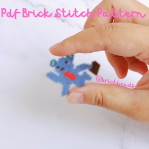 May include: A blue and pink pixelated bear with a red heart on its chest is being held by a hand. The text "@brickbeads" is visible on the hand.
