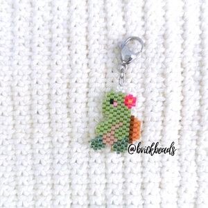 May include: A green and brown beaded turtle keychain with a pink flower on its head. The keychain is attached to a silver metal lobster clasp. The keychain is on a white knitted surface. @brickbeads