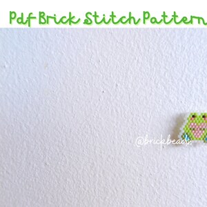 May include: A green frog bead sprite with pink cheeks and blue feet, created with perler beads. The bead sprite is on a white background. The text "@brickbeads" is in the lower left corner of the image.