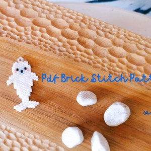 Pode incluir: Padrão de ponto de tijolo de foca em contas brancas sobre uma superfície de madeira com o texto "Pdf Brick Stitch Pattern" e o nome de usuário de mídia social "@brickbeads".