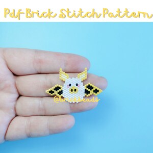 May include: A white pig with yellow and black wings, made with beads, on a blue background. The pig has a pink nose and eyes. The text "@brickheads" is visible below the pig.