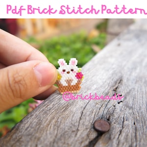 May include: A white bunny with pink ears and a pink flower sits in a brown basket with yellow accents. The bunny is holding a small white flower. The bunny is made of small beads and is a brick stitch pattern.