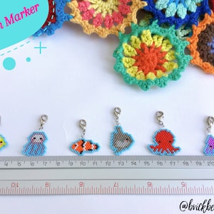 Puede incluir: Seis marcadores de punto de cuentas de colores con forma de criaturas marinas, incluyendo un pez amarillo, un pulpo morado, un pez payaso naranja y blanco, un tiburón gris, un pulpo rojo y un caballito de mar morado. Los marcadores cuelgan de cierres de langosta plateados y están alineados en una regla.