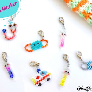 Op de afbeelding: Een set van zes kleurrijke kralen-steekmarkeerders met verschillende ontwerpen, waaronder een stethoscoop, een gezichtsmasker, een ambulance en een ijslolly. De steekmarkeerders zijn bevestigd aan zilveren clips. De tekst "Stitch Marker" is in roze op een blauwe achtergrond geschreven.