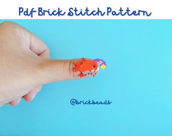 Puede incluir: Un pequeño pez naranja con un arcoíris en la parte superior, hecho con cuentas de colores. El pez está en el dedo de una persona. El fondo es una superficie azul claro. El texto "@brickbeads" está en la esquina inferior derecha de la imagen.