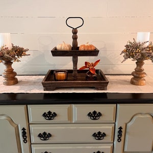 May include: A two-tiered wooden serving tray with black metal accents, decorated with fall-themed pumpkins and dried floral arrangements. The tray is displayed on a light brown dresser with black trim and decorative hardware.