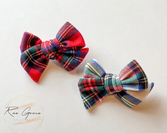 May include: Two plaid fabric bows in red, green, blue, and white. The bows are tied in a classic bow shape, perfect for hair accessories or crafting projects. The bows are on a white background.