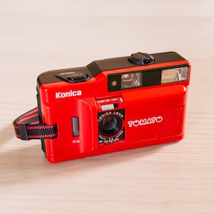 May include: A red Konica Tomato camera with a black top and a black and red striped wrist strap. The camera has a flash and a Konica lens. The word "TOMATO" is printed on the side.