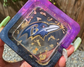 Op de afbeelding: Vierkante harslade met een Baltimore Ravens-logo. De lade heeft een zwart, paars en goud kleurenschema. Gouden vlokken zijn ingebed in de heldere hars. De lade wordt in een hand gehouden.