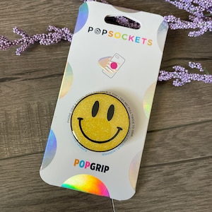 May include: Yellow smiley face phone grip with glitter. The grip is attached to a white card with the text "PopSockets" and "PopGrip".