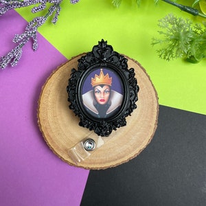 May include: Black and white Evil Queen character badge reel with a black ornate frame and a clear plastic retractable badge holder. The Evil Queen is wearing a purple dress and a gold crown.
