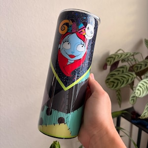 Puede incluir: Un vaso negro con purpurina con un dise&ntilde;o de la cara de Sally de Pesadilla antes de Navidad. El dise&ntilde;o incluye la cara, el pelo y el vestido de retazos de Sally. El vaso tiene una tapa negra.