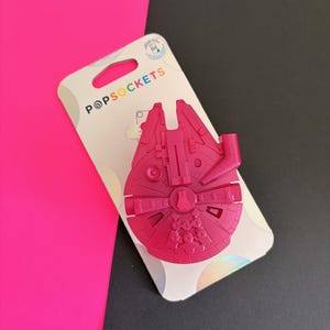 May include: A pink PopSockets phone grip shaped like the Millennium Falcon spaceship. The grip is attached to a white and iridescent backing card with the PopSockets logo. The background is a combination of black and bright pink.