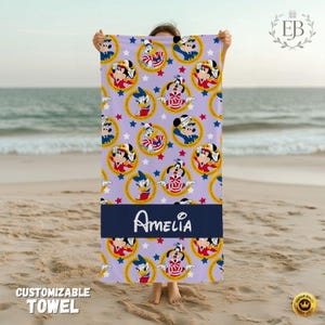 May include: A lavender beach towel featuring a pattern of cartoon characters in gold circles and red stars. A navy blue band displays the name "Amelia" in white script. The towel is held up on a sandy beach.
