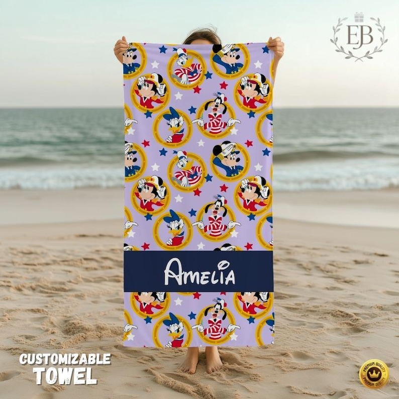 May include: A lavender beach towel featuring a pattern of cartoon characters in gold circles and red stars. A navy blue band displays the name "Amelia" in white script. The towel is held up on a sandy beach.