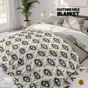 May include: A cream-colored blanket with a repeating pattern of blue and gold evil eye designs. The blanket is draped over a bed with gray pillows and a white sheet. The text "CUSTOMIZABLE BLANKET" is visible.