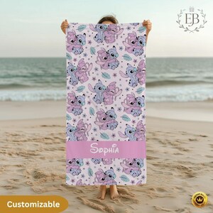 May include: A light pink beach towel featuring a repeating pattern of cartoon characters in shades of blue and pink. A pink band at the bottom displays the name "Sophia" in white script. The word "Customizable" is in a yellow oval at the bottom.