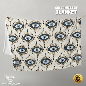 May include: A cream-colored blanket with a repeating pattern of blue and gold evil eye designs. The blanket is hanging from a string, with the text "CUSTOMIZABLE BLANKET" at the top. The blanket has a white backing.